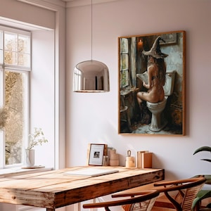 May include: A framed artwork shows a person wearing a witch hat, seated on a toilet, reading. The artwork is in a wooden frame and hangs on a white wall. A silver pendant light hangs above a wooden table. The background includes a window.