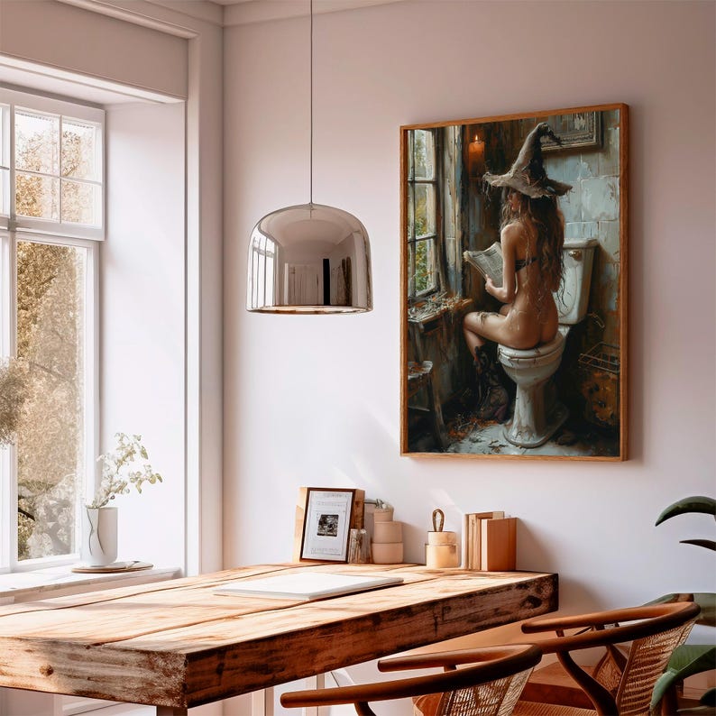 May include: A framed artwork shows a person wearing a witch hat, seated on a toilet, reading. The artwork is in a wooden frame and hangs on a white wall. A silver pendant light hangs above a wooden table. The background includes a window.
