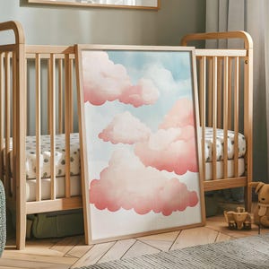 Op de afbeelding: Een ingelijste print van een aquarel van roze wolken tegen een blauwe lucht. De print is in een houten lijst en leunt tegen een houten wieg.
