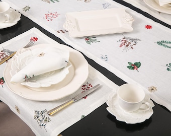 May include: A white table setting with a floral design. The setting includes a white tablecloth, plates, cups, saucers, and a rectangular serving dish. The tablecloth and napkins are decorated with red berries, green leaves, and other floral elements.