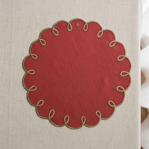 May include: A circular red placemat featuring a scalloped edge outlined with olive green embroidery. The center of the placemat is solid red fabric with a visible woven texture, and the decorative border consists of continuous looping stitches.