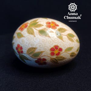 May include: A decorative egg with a cream-colored background, adorned with red flowers and green leaves. The egg has a floral pattern and the text "Anna Chumak Handmade".