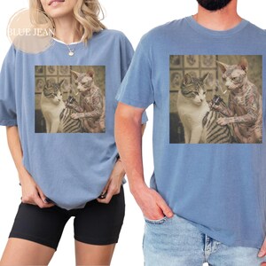 May include: Two light blue t-shirts featuring a square graphic of two cats. One is hairless and tattooed, holding a cigarette for the other. The text "BLUE JEAN" is in a circle in the upper left corner.