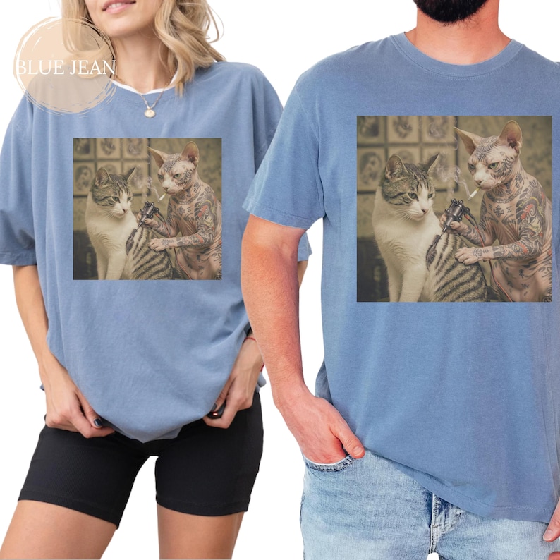 May include: Two light blue t-shirts featuring a square graphic of two cats. One is hairless and tattooed, holding a cigarette for the other. The text "BLUE JEAN" is in a circle in the upper left corner.