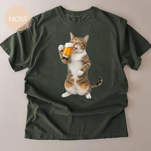 以下が含まれることがあります： ビールを飲んでいる猫のグラフィックが特徴の濃い緑色のTシャツ。猫は茶色、白、タン色で、舌を出しています。「MOSS」という文字がシャツの左上にプリントされています。