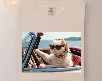 May include: Ivory t-shirt with a graphic of a white cat wearing sunglasses, a headscarf, and a pearl necklace, driving a red convertible. The cat is at the wheel, with a coastal backdrop of sea and hills.