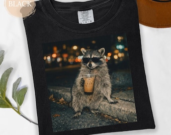 May include: Black t-shirt with a raccoon wearing sunglasses and holding an iced coffee. The raccoon is on a ledge with a blurred city background. The shirt has a Comfort Colors label.