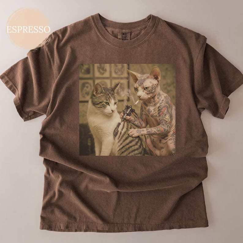 May include: Espresso-coloured t-shirt with a graphic of two cats, one tattooing the other. The tattooed cat is a hairless Sphynx, and the other is a tabby. The image has a vintage, slightly faded appearance.