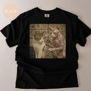 May include: Black t-shirt with a square image of two cats. One cat is tattooing the other cat. The tattooed cat is a hairless breed with full arm tattoos. The other cat is a tabby. The word "BLACK" is in a circle in the upper left corner.