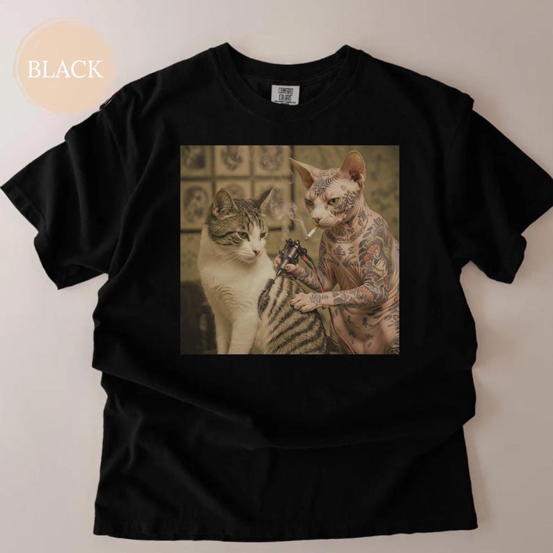 May include: Black t-shirt with a square image of two cats. One cat is tattooing the other cat. The tattooed cat is a hairless breed with full arm tattoos. The other cat is a tabby. The word "BLACK" is in a circle in the upper left corner.
