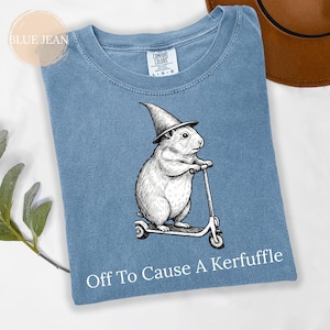 Könnte beinhalten: Blaues Jeans-T-Shirt mit einer Grafik eines Nagetiers, das einen Zaubererhut trägt und Roller fährt. Der Text "Off To Cause A Kerfuffle" ist unten aufgedruckt. Das T-Shirt hat einen Rundhalsausschnitt und kurze Ärmel.