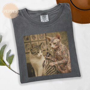 May include: Dark grey Comfort Colors t-shirt with a humorous cat tattoo design. One cat is tattooing another cat, who is smoking a cigarette. The shirt has a crew neck and short sleeves.