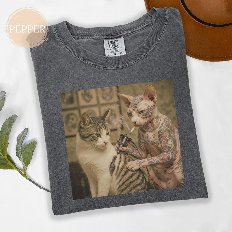 May include: Dark grey Comfort Colors t-shirt with a humorous cat tattoo design. One cat is tattooing another cat, who is smoking a cigarette. The shirt has a crew neck and short sleeves.