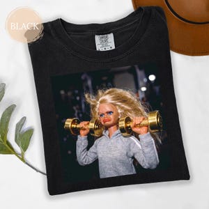 Könnte beinhalten: Schwarzes Comfort Colors T-Shirt mit einer Puppe, die goldene Hanteln hebt. Die Puppe trägt einen grauen Kapuzenpullover mit weißen Streifen. Das Shirt hat das Wort "BLACK" in einem Kreis oben links.