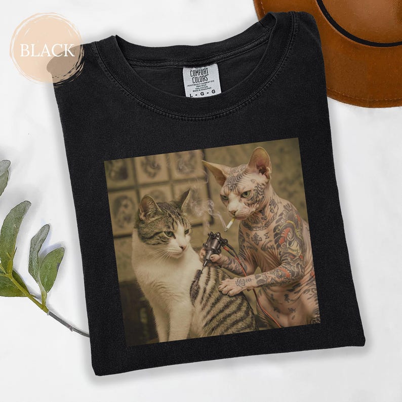 May include: Black Comfort Colors t-shirt with a humorous graphic of two cats. A tattooed Sphynx cat is tattooing another cat. The image includes a lit cigarette and a tattoo gun.