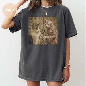 May include: Dark grey t-shirt with a square graphic depicting two cats. One cat tattoos the other. The tattooed cat is a hairless Sphynx. The other cat is a tabby. The shirt has a loose fit.
