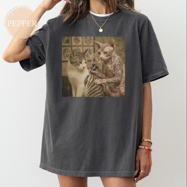 May include: Dark grey t-shirt with a square graphic depicting two cats. One cat tattoos the other. The tattooed cat is a hairless Sphynx. The other cat is a tabby. The shirt has a loose fit.