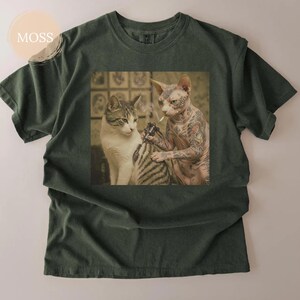 May include: Dark green t-shirt with a graphic of two cats. A tattooed Sphynx cat is tattooing a tabby cat, while smoking a cigarette. The tee has a crew neck and short sleeves. The image is printed on the front.