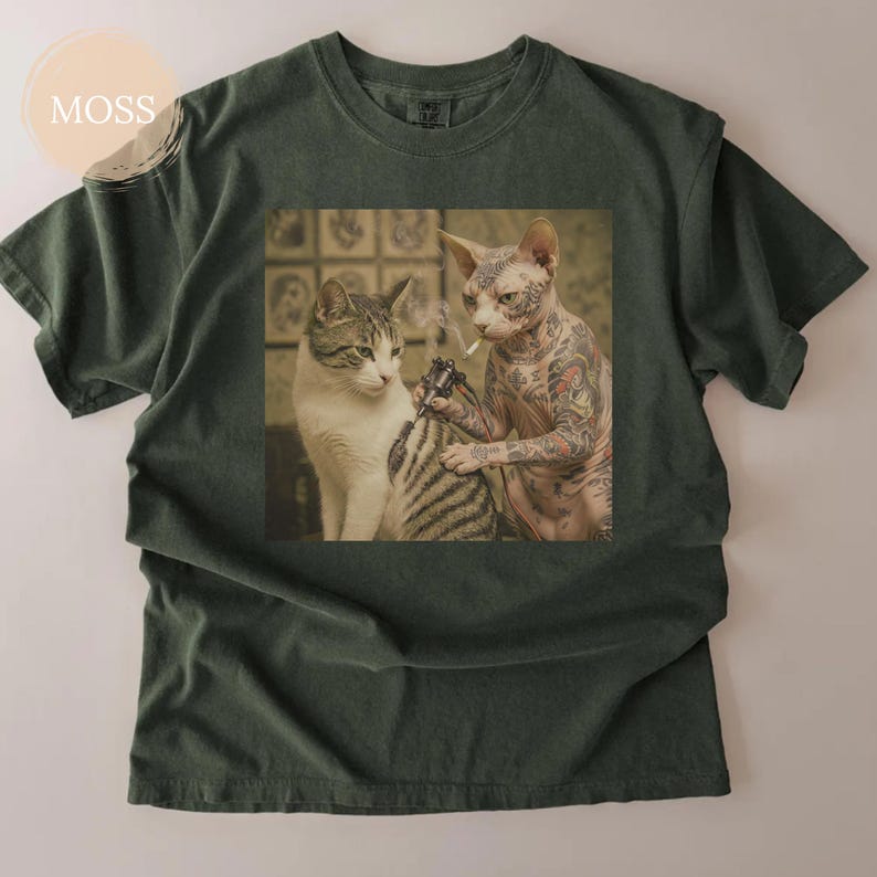 May include: Dark green t-shirt with a graphic of two cats. A tattooed Sphynx cat is tattooing a tabby cat, while smoking a cigarette. The tee has a crew neck and short sleeves. The image is printed on the front.