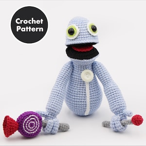 Könnte beinhalten: Hellblaue Häkelpuppe mit großen grünen und schwarzen Augen, einem schwarzen Mund und einem roten Innenraum. Die Puppe hält einen lila und roten Lutscher in der einen Hand und einen roten Ball in der anderen. Der Text "Crochet Pattern" befindet sich in einem schwarzen Kreis.