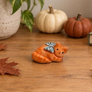 May include: A small ceramic figurine of a curled-up orange tabby cat with a light blue bow on its back. The cat has a smooth, glossy finish, pointed ears, and two small black dots for eyes, resting in a resting position on a wooden surface.