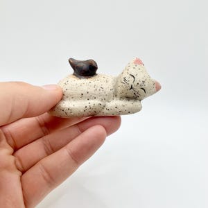May include: A ceramic cat figurine in a sleeping pose. The cat is off-white with black speckles, pink accents on the ears and nose, and closed eyes. A small, dark brown object rests on its back.