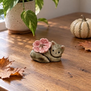 May include: A small, smooth, grey ceramic figurine of a resting cat with a large, textured, light pink flower attached to its back. The cat has simple, dark, circular eyes and a pointed ear, featuring a glossy finish that reflects light.