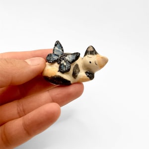 May include: A small, ceramic figurine of a sleeping cow with black and white spots, a tan face, and a blue butterfly perched on its back. The figurine is held in a hand against a white background.