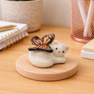 May include: A white ceramic cat figurine with a butterfly on its back, resting on a round wooden base. The butterfly has orange, black, and brown wings. The figurine is on a wooden desk.