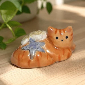 May include: A small ceramic figurine of an orange tabby cat with dark orange stripes, two black eyes, and a black nose. A white seashell and a light blue starfish are attached to the cat's back. The cat is in a resting, lying-down position.