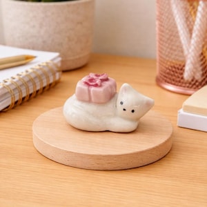 May include: A white ceramic cat figurine with a pink square-shaped object on its back, resting on a round wooden base. The cat has black eyes and is lying down. The figurine is on a wooden desk.