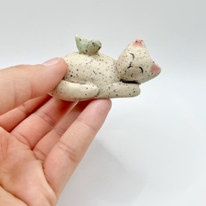 May include: A ceramic cat figurine in a relaxed pose, with a small bird perched on its back. The cat is off-white with black speckles, pink accents on the ears and nose, and closed eyes. The bird is light green. The figurine is held in a hand.