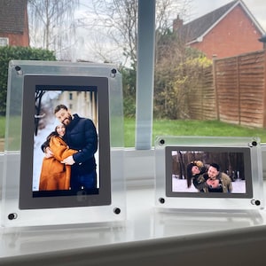 May include: Two clear acrylic photo frames displaying colour photographs of a couple. The frames are rectangular with rounded corners and black screw accents. The background shows a window with a view of trees and houses.