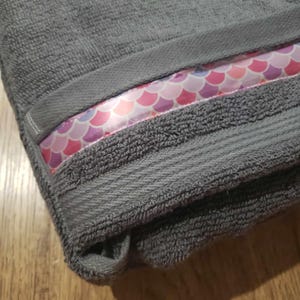 Puede incluir: Una toalla gris doblada con una banda decorativa con un patrón de escamas de sirena en rosa, morado y blanco. La toalla tiene un aspecto texturizado y un borde doblado.