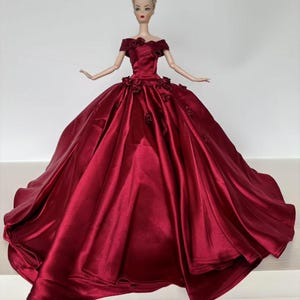 May include: A doll wearing a formal, floor-length gown in a deep red hue. The off-the-shoulder dress features rose accents on the bodice and skirt. The doll has blonde hair and is posed with arms outstretched.