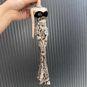 Peut inclure: Une tenue de poupée comprenant un haut court en dentelle noire et une jupe à imprimé peau de serpent. La jupe est une conception ajustée et longue. Un collier en argent est également inclus. La tenue est conçue pour une poupée.