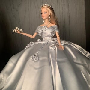 May include: A silver satin ball gown with a sweetheart neckline and a fitted bodice. The gown is adorned with large, white fabric roses and a silver tiara. The doll is wearing a pearl necklace and silver earrings.