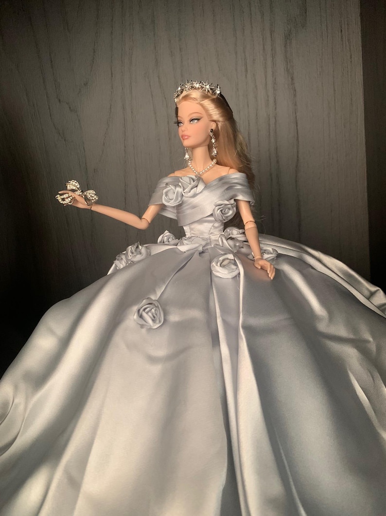 May include: A silver satin ball gown with a sweetheart neckline and a fitted bodice. The gown is adorned with large, white fabric roses and a silver tiara. The doll is wearing a pearl necklace and silver earrings.