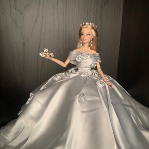 May include: A silver satin ball gown with a white rose design and a silver tiara on a doll.