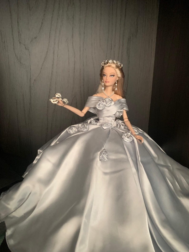 May include: A silver satin ball gown with a white rose design and a silver tiara on a doll.