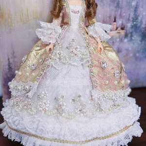 Puede incluir: Una muñeca con un vestido blanco y dorado con ribetes de encaje y adornos de perlas. El vestido tiene un corpiño rosa y dorado con cintura ajustada y falda completa.