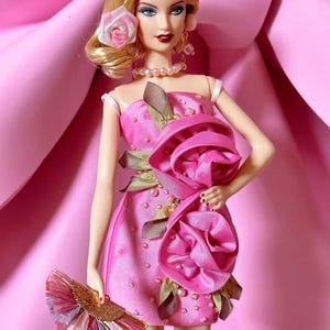 Puede incluir: Un vestido rosa con una gran rosa rosa en el lateral. El vestido está decorado con pequeñas cuentas rosas y hojas verdes. La muñeca lleva un collar de perlas y pendientes.