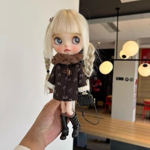 Op de afbeelding: Een pop met blonde vlechten, gekleed in een bruine jas met witte rand, een bruine sjaal en zwarte laarzen. De pop houdt een kleine zwarte handtas met een zilveren ketting vast. De pop heeft grote blauwe ogen en roze wangen.