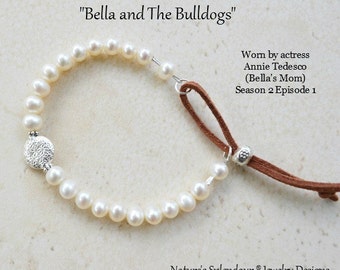 May include: A white pearl bracelet with a silver charm and a brown leather cord. The bracelet is worn by Annie Tedesco, who plays Bella's mom in the Nickelodeon show "Bella and The Bulldogs".