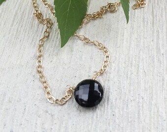 May include: A gold-colored chain necklace with a faceted, round, black gemstone pendant. The necklace is displayed on a light-colored surface with green leaves in the background. The chain has a delicate, linked design.