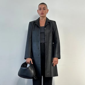 May include: A black leather coat with a button-up front, paired with black pants and a matching black handbag. The coat has a classic, tailored design. The outfit is completed with black sandals.