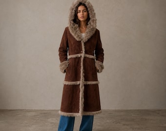 Peut inclure: Un long manteau marron foncé doté d'une capuche, de poignets et d'une bordure avant garnis de fausse fourrure marron clair. Le manteau présente une fermeture à un bouton à la taille et des coutures horizontales sur le torse, porté avec un jean bleu.