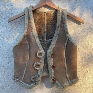 Pode incluir: Um colete de camurça marrom com forro de pele de carneiro e quatro botões marrons. O colete tem um decote em V e é feito de um material macio e luxuoso.