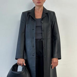 May include: A black leather coat with a button-up front, worn over a gray top and black pants. The coat is knee-length and has a classic lapel collar. A black handbag is held in the left hand. The background is a plain white wall.