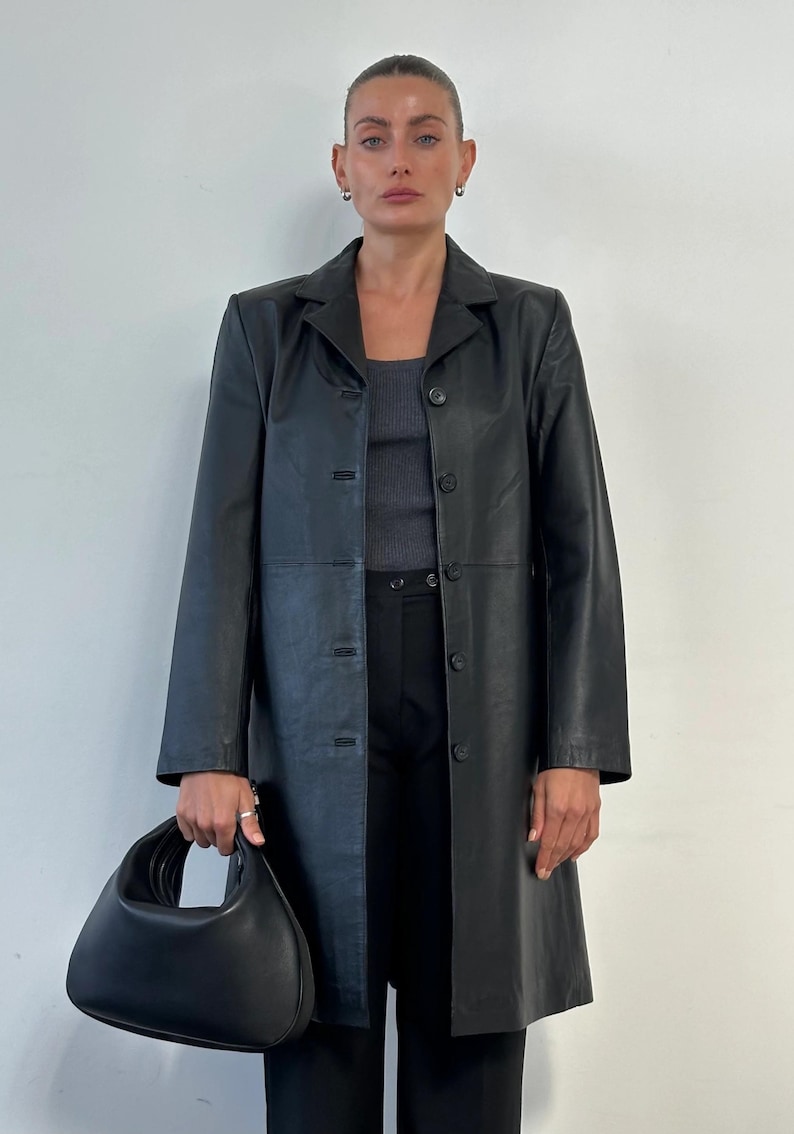 May include: A black leather coat with a button-up front, worn over a gray top and black pants. The coat is knee-length and has a classic lapel collar. A black handbag is held in the left hand. The background is a plain white wall.